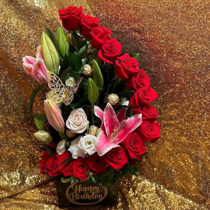 A box arrangement on a gold sequin backdrop: a sweeping arc of red roses, pink stargazer lilies, a cream rose and white lisianthus, foil-wrapped Ferrero Rocher chocolates on picks and a gold plaque on the box reading Happy Birthday.