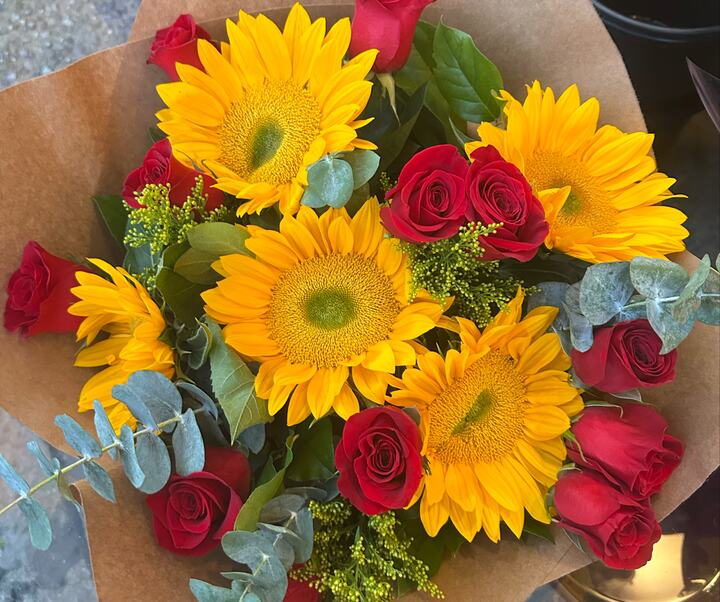 A bouquet seen from above: sunflowers and red roses with green foliage, wrapped in brown kraft paper.