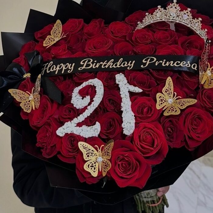 A wide bouquet of red roses fanned in black paper with a pearl tiara at the top, gold butterflies, a black bow, a sash reading Happy Birthday Princesa, and the number 21 picked out in white gypsophila across the centre.