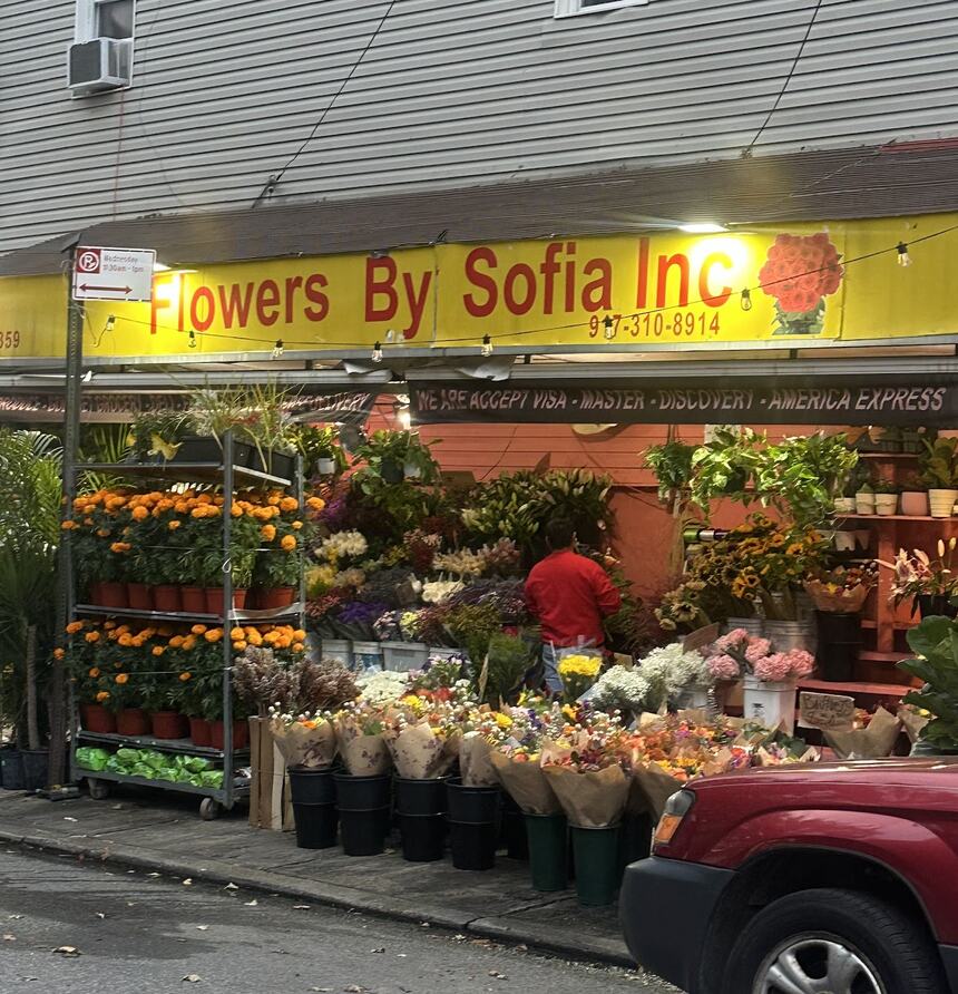 The Flowers by Sofia stand on Graham Ave: a lit yellow sign reading Flowers By Sofia Inc, racks of orange marigolds, and wrapped bouquets standing in buckets along the sidewalk.