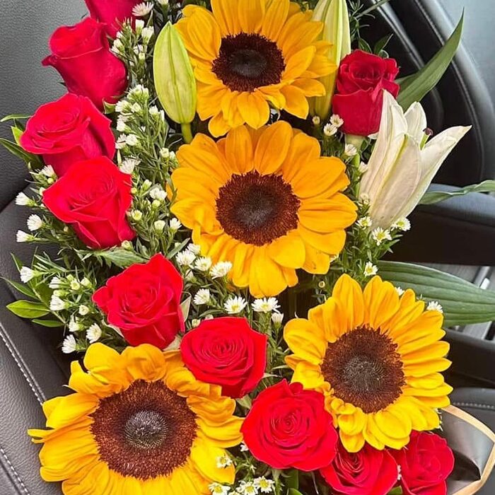 A tall arrangement in a black box: four large sunflowers down the centre, a column of red roses along one side, white lily buds, waxflower and daisies filling the gaps.